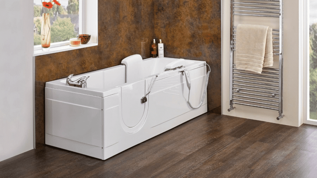 A close-up view of the white 'Montana' walk-in bath featuring a rectangular, wide-access door with a side panel. The interior of the full-length tub shows an integrated high seat, chrome grab handle, and an electric bidet wash wand with a control unit. Positioned in a modern bathroom with dark, textured copper wall tiles, this bath highlights premium features designed for safe, independent bathing.