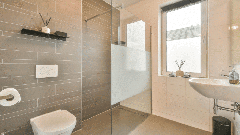 A clean, modern, and minimalistic accessible wet room featuring a level-access shower area with a linear floor drain and a walk-in glass panel with a central frosted privacy band. To the left is a wall-mounted toilet with large grey textured tiles, while the right features a simple sink and white tiles next to a large window.