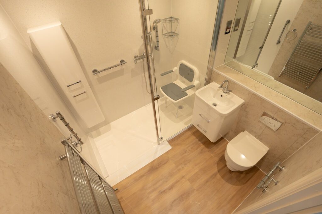 Maya's finished bathroom from above: low-level walk-in shower with fold-down seat and grab rails, vanity basin and wall-hung toilet.